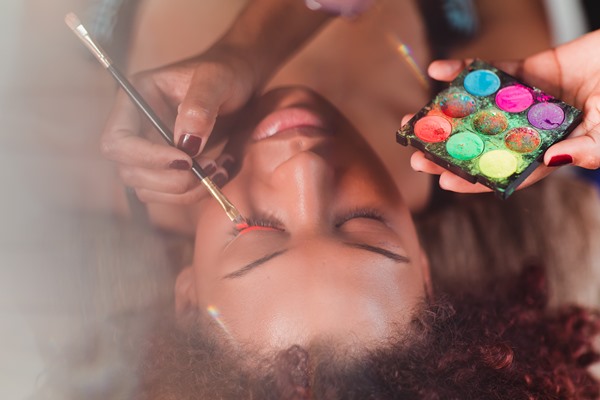 Mulher negra, com cabelo cacheado, sendo maquiada por uma mulher para uma competição. A modelo participou do concurso de beleza Top Cufa DF