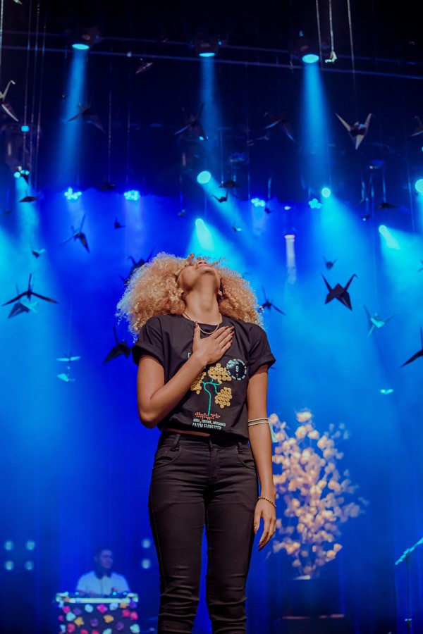 Mulher jovem e negre, com cabelo cacheado louro, e camiseta e calças pretos. Ela está em cima de um palco fazendo o movimento de agradecimento. O evento é o concurso de beleza Top Cufa DF.