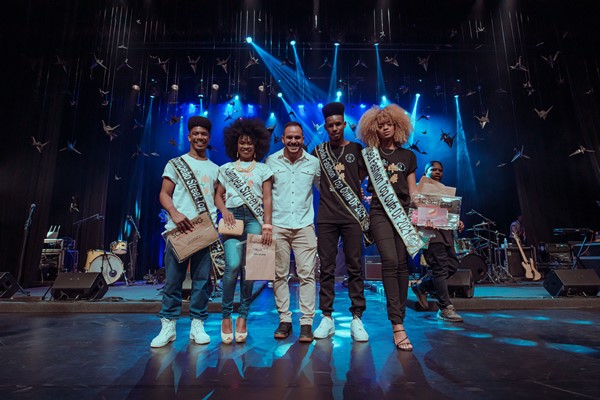 Finalistas do concurso de beleza Top Cufa 2021 no palco e o apresentador da competição. Da esquerda para direita estão: um homem negro com cabelo black, uma mulher negra com cabelo cacheado preto, um homem negro careca, um homem negro com cabelo black e uma mulher negra com cabelo cacheado louro.