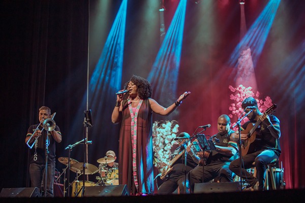 Mulher negra com cabelo cacheado médio preto. Ela está em um palco com luzes azuis e roxas e segura o microfone, cantando. No fundo é possível ver outras pessoas tocando instrumentos, como bateria e violão