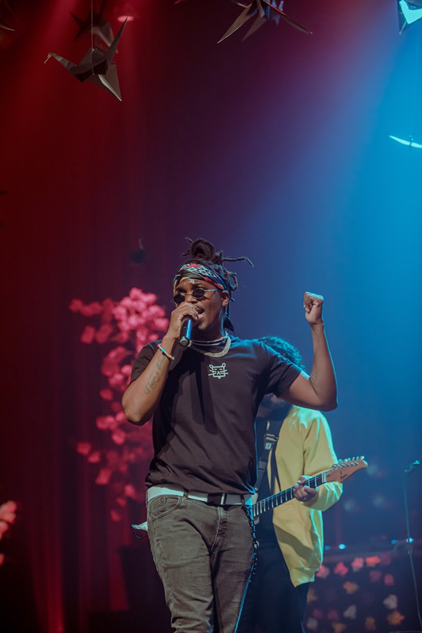 Homem negro com tranças médias amarradas e uma bandana. Ele está em um palco com luzes azuis e roxas e segura o microfone, cantando. No fundo é possível ver outra pessoa tocando violão