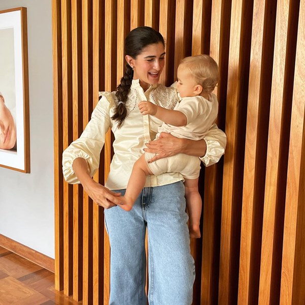 Mulher branca, com cabelos castanhos arrumados em tranças, segurando o próprio filho, um bebê de menos de um ano. Ela usa uma camisa de botões brancas, de seda, com calça jeans de lavagem clara.