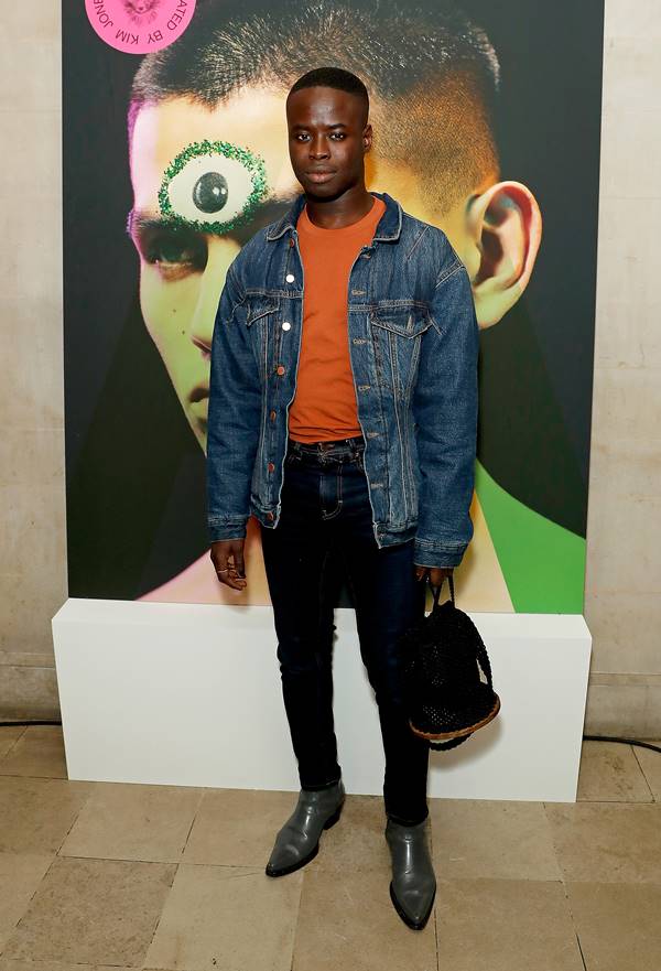 Homem negro usa camiseta laranja, jaqueta jeans, calça preta e segura bolsa na mão direita