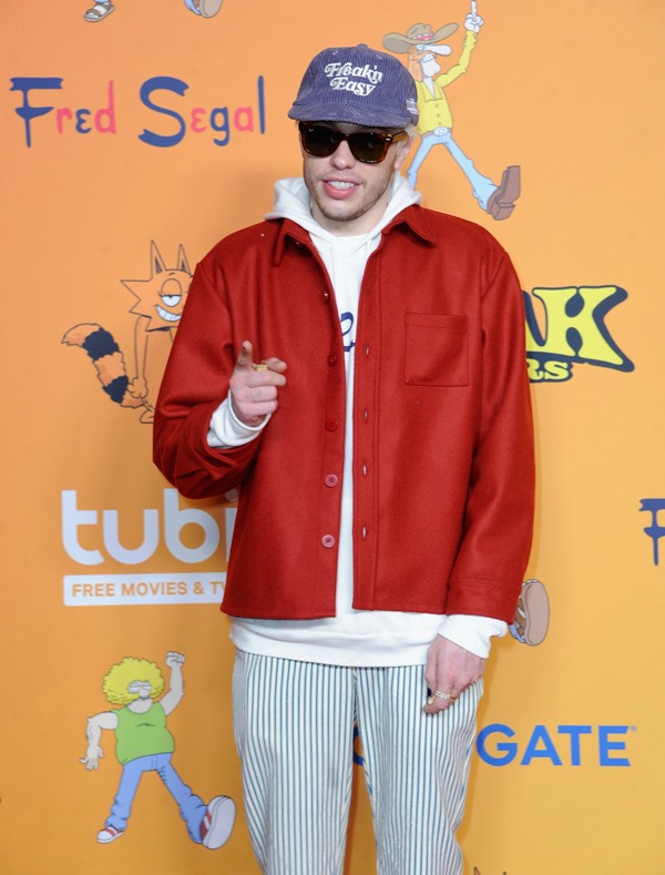 O comediante norte-americano Pete Davidson em entrada de evento. Ele é um homem branco, com cabelo descolorido, e está posando para foto. Ele veste um moletom branco, uma jaqueta de couro vermelha/marrom, uma calça moletom cinza, um óculos escuros preto e boné no tecido jeans.