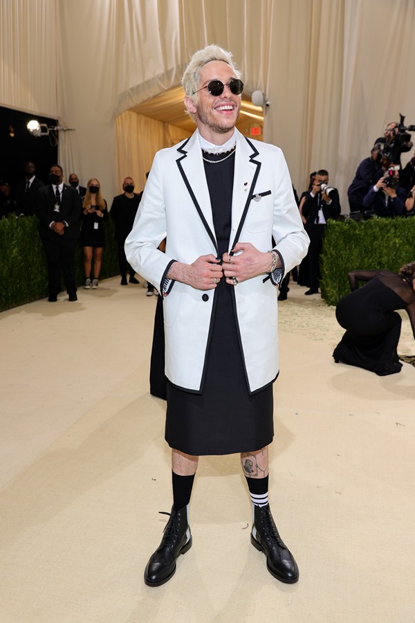 O comediante norte-americano Pete Davidson no tapete vermelho do evento MET Gala de 2021. Ele é um homem branco, com cabelo louro descolorido, e está posando para foto. Ele usa um vestido preto com gola branca, blazer branco com detalhes pretos, meia preta e sapato estilo oxford preto. Usa, ainda, um óculos escuros pretos.