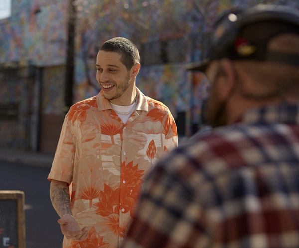 O comediante norte-americano Pete Davidson em campanha de divulgação da nova coleção masculina da marca H&M. Ele usa uma camisa colorida de botões, com manga curta, e estampa de folhas laranja; uma camiseta branca por baixo; corrente prateada e uma bermuda lilás. 