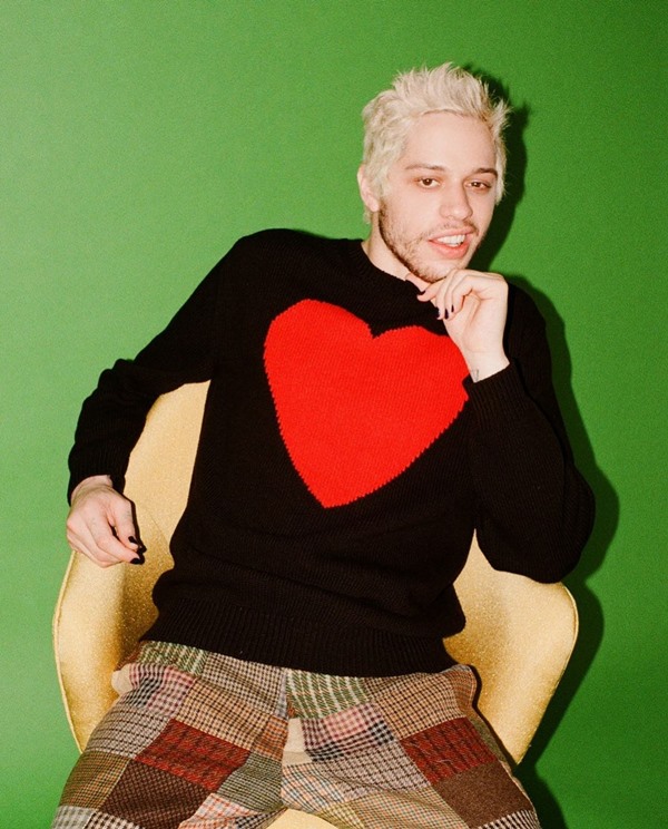 O comediante norte-americano Pete Davidson em campanha de divulgação da nova coleção da marca Rowing Blazer. Ele é um homem branco, com cabelo descolorido, e está posando para foto. Usa um casaco de tricô preto com um coração vermelho e grande no centro, e uma calça xadrez bege. Está sentado em uma poltrona na cor amarela clara.