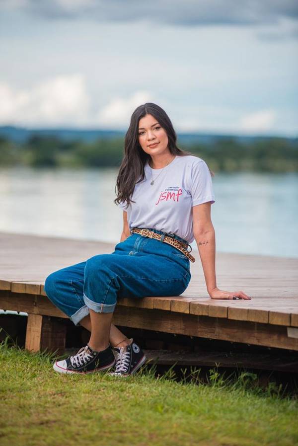 Mulher branca sentada em deck, usando calça jeans e camiseta branca