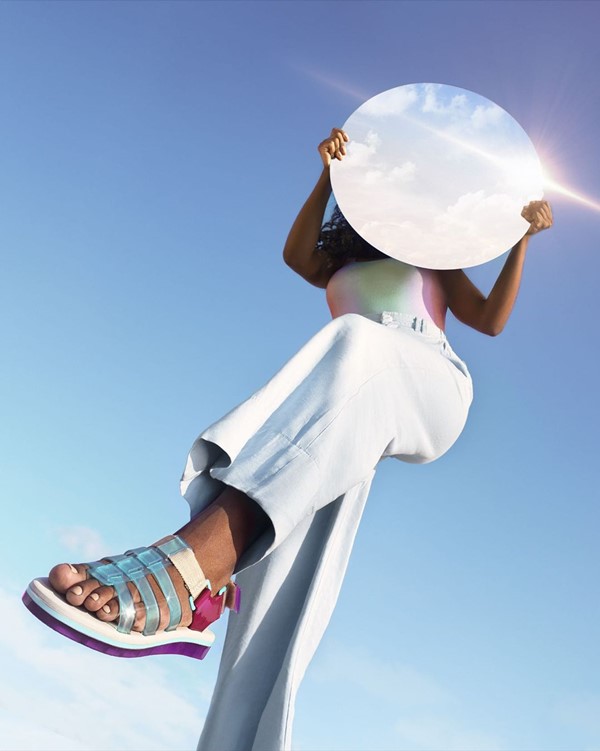 Mulher negra com cabelos cumpridos posando para foto ao ar livre. Ela segura um espelho redondo, que reflete o céu azul e as nuvens brancas. Ela veste uma calça jeans clara, um body colorido e uma sandália da Melissa nas cores branco, azul e roxo.