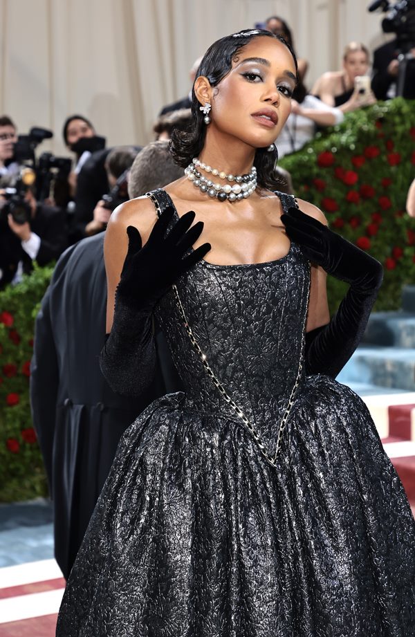 A atriz Laura Harrier, uma mulher negra de cabelos pretos, no tapete vermelho do Met Gala de 2022. Ela veste um vestido preto brilhante, luvas também pretas e colares de pérolas pretas e cinzas. O vestido faz referência às roupas do século 18.