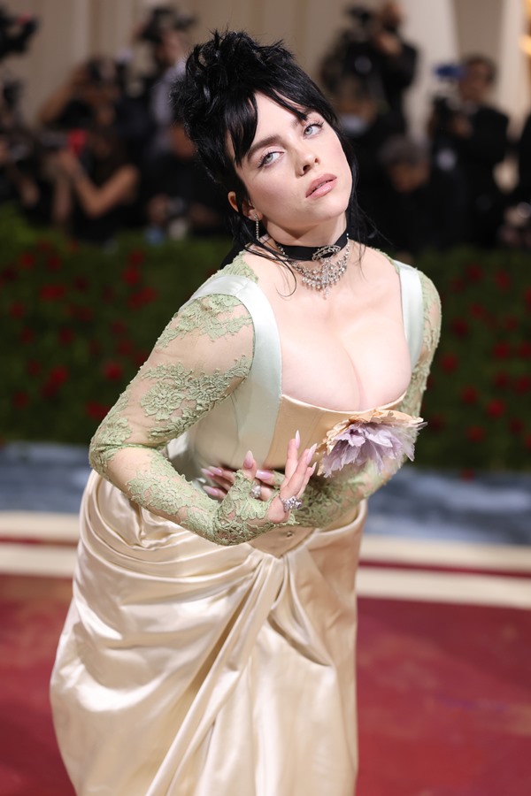 A cantora Billie Eilish no tapete vermelha do Met Gala de 2022. Ela, uma mulher branca de olhos azuis e cabelos pretos, usa um vestido bege de seda que imita as peças usadas no século 18. Usa, ainda, uma gargantilha preta e correntes de prata.