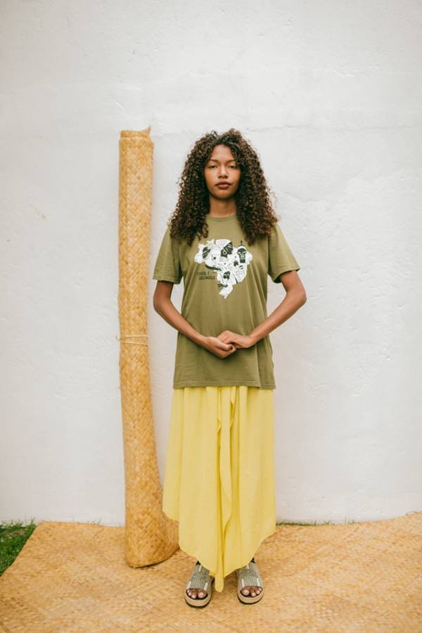 Mulher negra posa em frente a parede branca usando camiseta verde e calça amarela