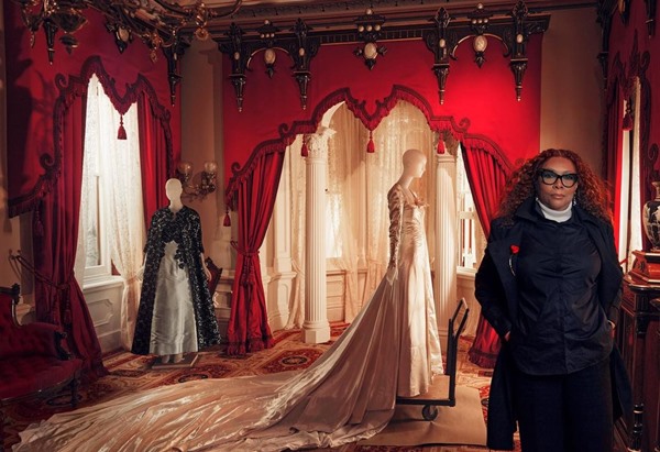 A diretora de cinema Julie Dash, uma mulher negra com cabelos grandes cacheados, posando para foto em uma das salas da nova exposição do Costume Institute, no Metropolitan Musem of Art de Nova York. No fundo é possível ver manequins vestindo roupas do século 18