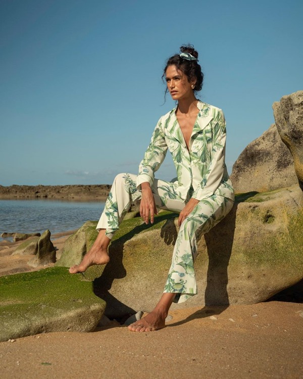 Modelo negra, com cabelos ondulados amarrados, posando para foto em uma praia com pijama de seda. O conjunto de calça e camisa é na cor branca, com estampa de plantas verdes, e no cabelo ela está com um enfeite na mesma estampa. As peças são da marca Chris Gontijo.