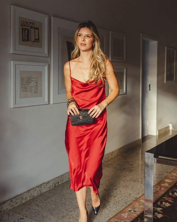 Mulher branca e loura, chmada Duda Portela Amorim, posando para foto na sala de casa com um vestido vermelho seda com alças finas. A peça é da marca mineira Chris Gonijo. Ela usa um salto scarpin preto e uma bolsa tamém preta, da marca Chanel.