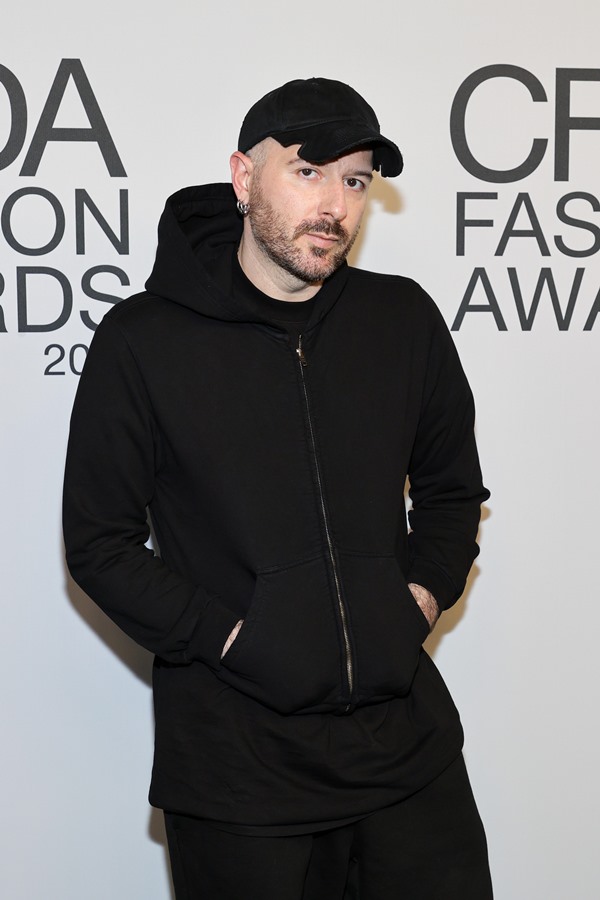 Homem branco, com a cabeça raspada e bigode, posando para foto em um evento. Seu nome é Demna Gvasalia e ele é o estilista da marca Balenciaga. Na foto, usa camiseta, casaco, calça e boné, tudo preto.