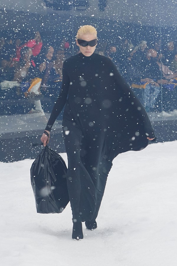 Mulher branca, com cabelo louro curto penteado para trás, desfilando na passarela da marca Balenciaga. O cenário imita uma rua durante o inverno, com neve no chão e caindo "do céu". Ela usa vestido longo com manga, sapato e óculos, tudo preto.