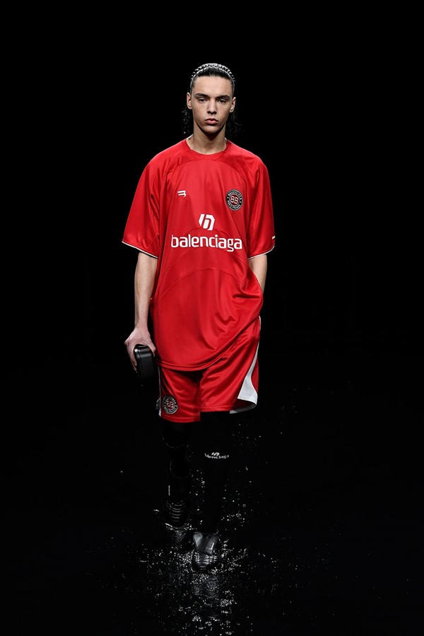 Homem branco, com cabelo preto curto penteado para trás, desfilando na passarela da marca Balenciaga. O cenário é todo preto e não é possível ver nada. Ele usa camiseta e shorts vermelho que lembram o uniforme de jogadores de time de futebol. 