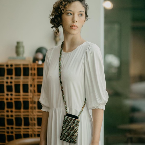 Mulher branca com cabelo cacheado curto posando para foto na varanda de uma casa. Ela usa um vestido branco de mangas cumpridas e uma bolsinha de crochê pequena, só para o celular.