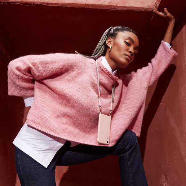 Mulher negra com tranças louras amarradas em um rabo de cavalo. Ela posa para foto em um fundo escuro, vermelho. Veste uma camisa branca e, por cima, um casaco rosa peludinho. Usa também uma capinha rosa para celular com corda que fica pendurada no pescoço.