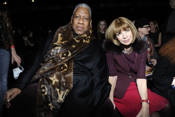 Os jornalistas Andre Leon Talley e Anna Wintour sentados na primeira fila de um desfile de moda. Ambos trabalham na revista Vogue. Andre é um homem negro e mais velho, com cabelo curto e branco. Usa uma roupa toda preta e, por cima, uma capa estilo toalha da marca Louis Vuitton. Já Anna é uma mulher de meia idade branca, com cabelo chanel louro, e usa um casaco vinho com saia vermelha.