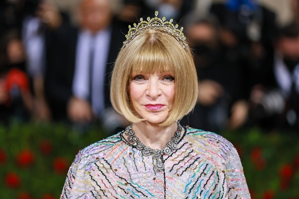 Anna Wintour, jornalista de moda e editora da revista Vogue, posando para foto no MET Gala de 2022. Ela é uma mulher idosa branca, com cabelo em corte chanel louro, e uma franja que cobre a testa. Veste uma roupa rosa brilhante, com detalhes de pluma e, na cabeça, uma tiara de diamantes.