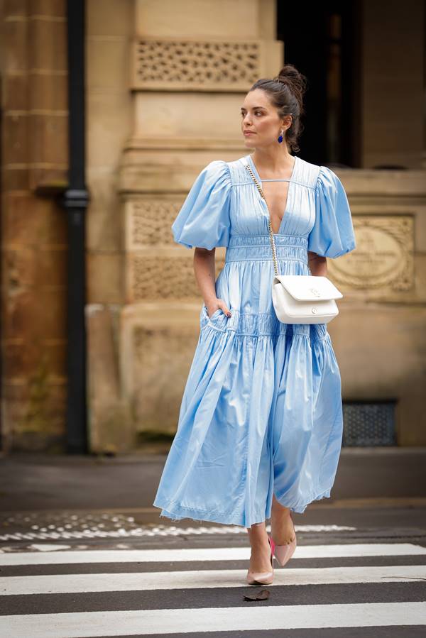 No street style, mulher usa vestido azul claro com mangas bufantes laterais e franzidos frontais, além de bolsa branca lateral