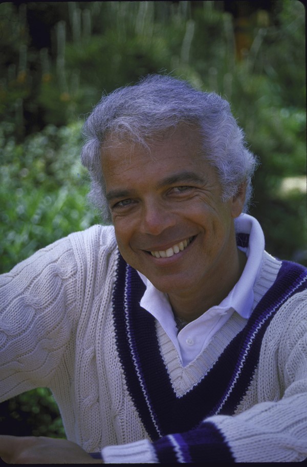 O estilista Ralph Laurne, um homem branco de cabelo grisalho, posando para foto em 1986. Ele usa roupas da própria marca: uma camisa polo branca e, por cima, um suéter bege com detalhes em azul marinho.