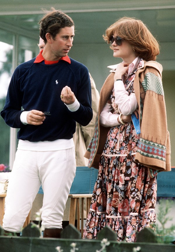 O príncipe Charles, ao lado de Lady Di, em 1977 em um jogo de polo equestre. Ele, um homem branco de cabelos castanhos, usa calça branca, camisa polo vermelha e suéter azul marinho. Ela é uma mulher branca com cabelos curtos e louros escuros. Veste uma camisa rosa, um vestido florido e um sobretudo caramelo, e usa óculos escuros.