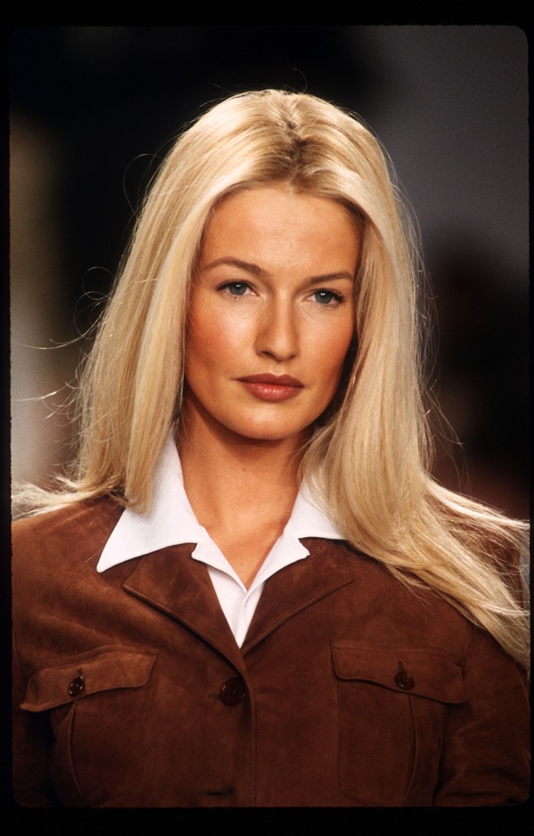 A modelo Karen Mulder, uma mulher branca, com cabelos louros lisos e longos, desfilando na passarela da marca Ralph Lauren. A foto é de 1996. Ela usa uma camisa polo branca e, por cima, uma jaqueta marrom de camurça. 