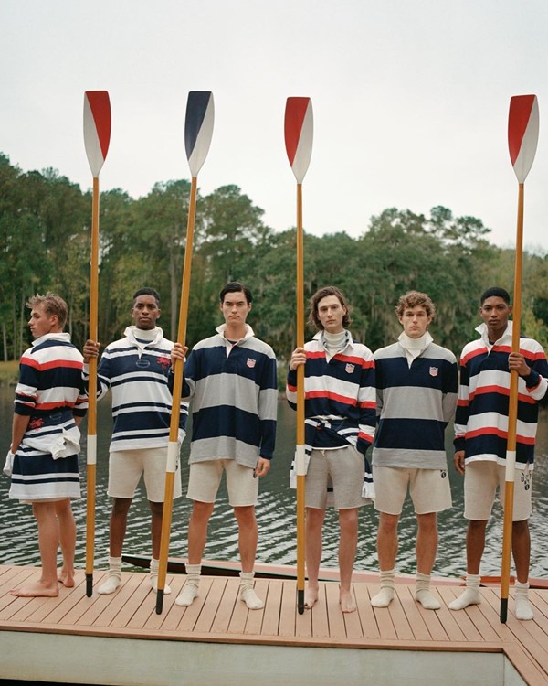 Atletas do remo posando para foto em um deque de madeira, com o rio ao fundo. Todos veste roupas da Ralph Lauren: camisas polos listradas e bermudas beges. 