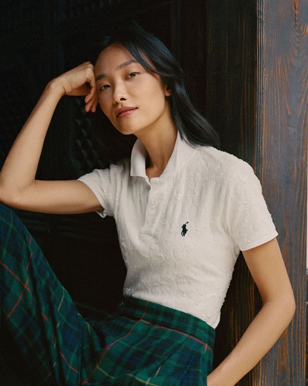 Modelo asiática, de pele clara e cabelos pretos ondulados, posando para foto em campanha da marca Ralph Lauren. Ela usa uma camisa polo branca, com textura de renda, e uma calça xadrez azul marinho e verde.