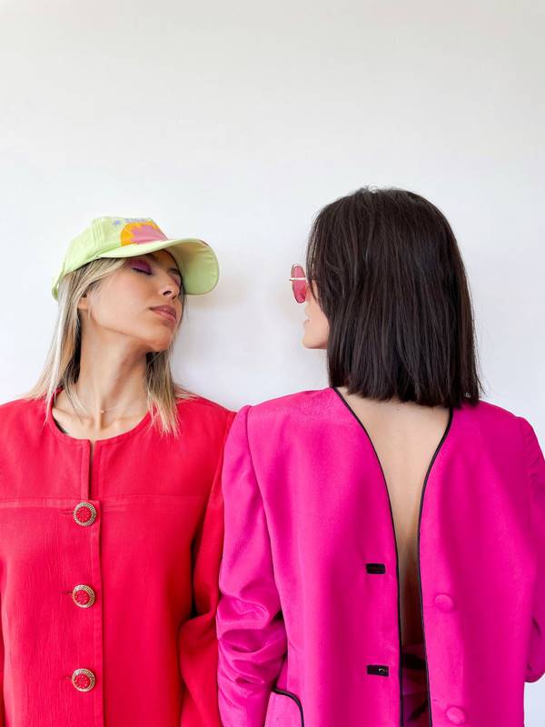 duas modelos se olham. um está usando um blazer rosa e a outra um blazer vermelho e usa também um bonê