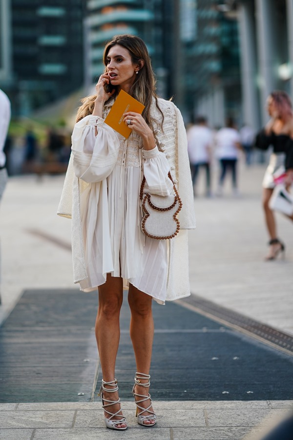 Mulher branca, com cabelos longos e ondulados, usa brincos, um mini vestido bordado plissado na cor creme com mangas bufantes, um casaco bordado na cor creme, uma bolsa cor creme decorada com pérolas marrons, sandálias gladiadoras de salto alto na cor cinza claro, fora do desfile Alberta Ferretti durante a Semana de Moda de Milão.