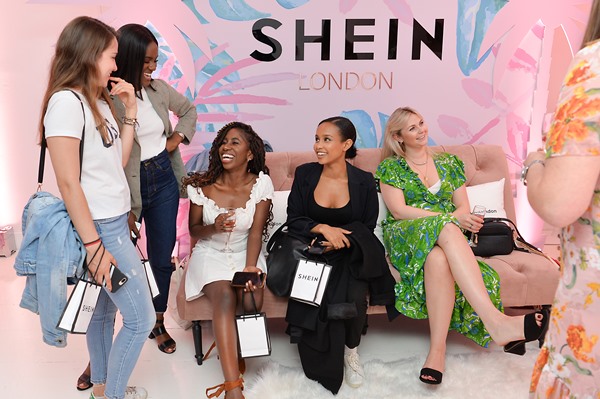 Evento da marca Shein. Na foto é possível ver um sofã e uma parede com desenhos e o logotipo da empresa, além de várias mulheres contratadas para divulgar a Shein. São mulheres magras, algumas brancas, outras morenas e outras negras.