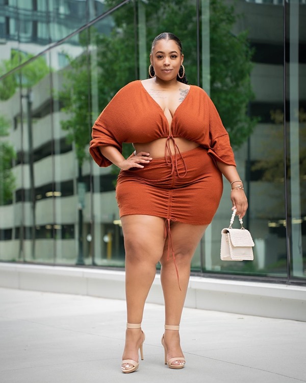 Modelo plus size posando para foto com roupas da Shein. É uma mulher negra com cabelo amarrado e brincos de argola dourada que veste um top e uma saia, tudo marrom, e da Shein. Usa ainda uma sandália de salto fino e uma bolsa pequena e branca.