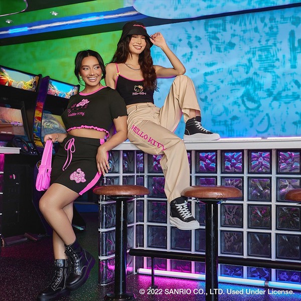 Campanha de divulgação da coleção da marca Shein com a boneca Hello Kitty. Na foto, duas mulheres posam para foto em um balcão de bar com roupas do lançamento. A primeira é uma mulher negra que usa shorts e top preto. A segunda e branca e usa top e chápeu preto e uma calça bege.