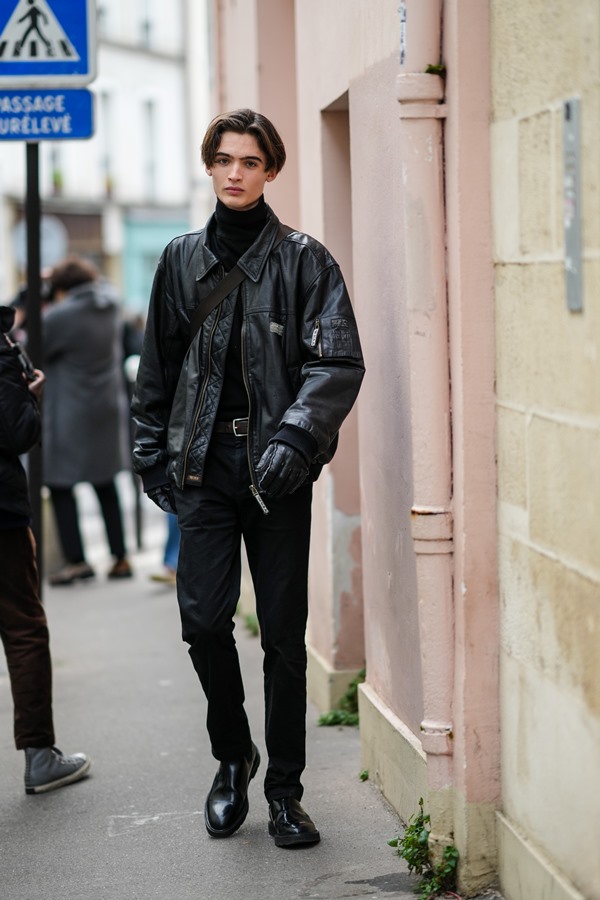 Homem branco e jovem, com cabelo liso e castanho, nas ruas de Paris durante a Semana de Moda. Ele usa um pulôver preto de gola alta, uma jaqueta preta de couro brilhante, um cinto de couro preto brilhante, luvas de couro preto brilhante, calça preta, botas pretas de couro brilhante.