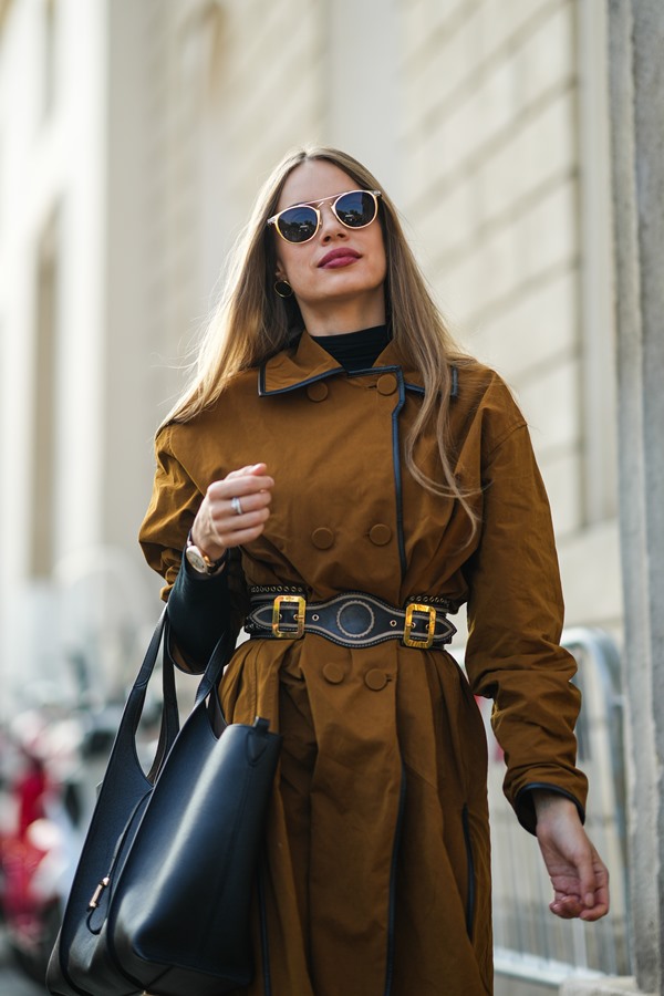 Mulher branca com cabelos longos, louros e lisos. Ela usa óculos de sol pretos, um pulôver preto de gola rulê, um casaco longo oversized com botões de camelo, um cinto de couro preto brilhante pregado/cravejado, uma bolsa grande de couro preto brilhante da Tod's, um cinto de couro preto, anéis de prata, fora do Desfile de moda de Tod's durante a Semana de Moda de Milão