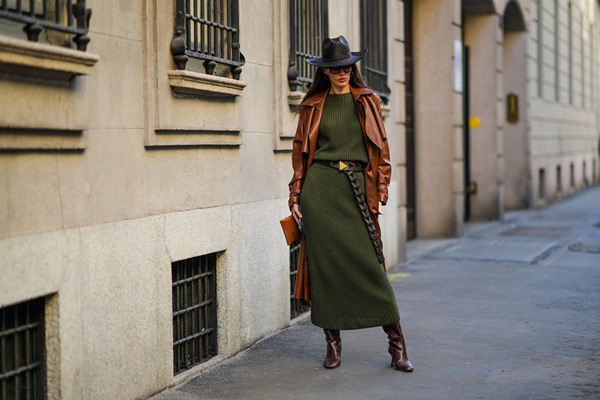 Mulher branca, com cabelos castanhos lisos, posando para foto na rua. Ela usa um chapéu de cowboy de vime trançado preto, óculos de sol marrons, brincos marrons, um vestido longo de lã com nervuras verde escuro, um casaco longo de couro brilhante de camelo, um cinto preto com padrão recortado, uma bolsa de couro brilhante de camelo, marrom escuro brilhante botas de salto alto com estampa de crocodilo em couro.