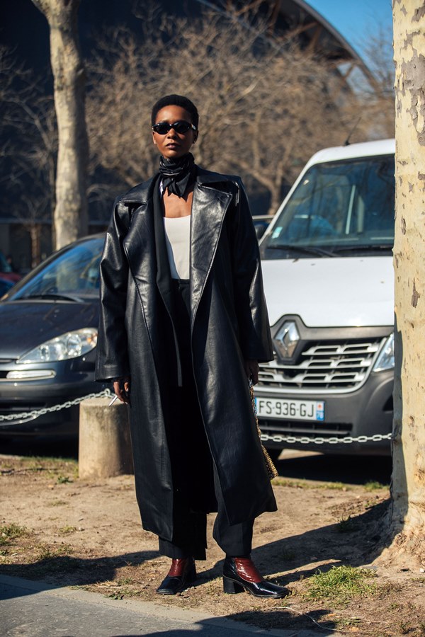 Mulher modelo negra de cabelo curto nas ruas de Paris durante a Semana de Moda. Ela usa óculos de sol pretos, um lenço preto no pescoço, trench coat de couro preto comprido, regata branca, calça preta e botas quadradas de látex pretas e marrons.