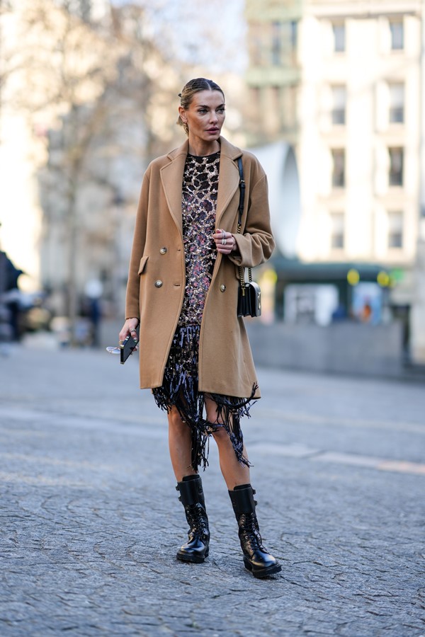 Mulher branca, com cabelo liso e louro, preso em um rabo de cavalo. Ela está andando nas ruas de Paris e veste brincos de ouro, um vestido bege com estampa de leopardo preto e lantejoulas, um casaco longo marrom, uma bolsa de couro preta brilhante, anéis de prata e strass, botas de couro preto brilhante mês/cadarços.