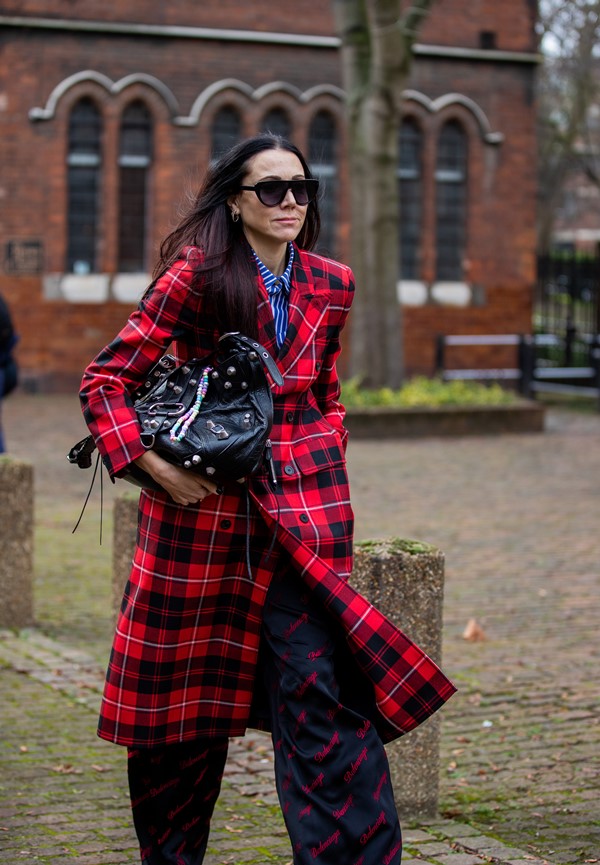 Mulher branca e de meia idade, com cabelos lisos, cumpridos e castanhos. Ela está andando pelas de Londres com um casaco xadrez vermelho, uma calça preta, óculos escuros e uma bolsa preta da marca Balenciaga.