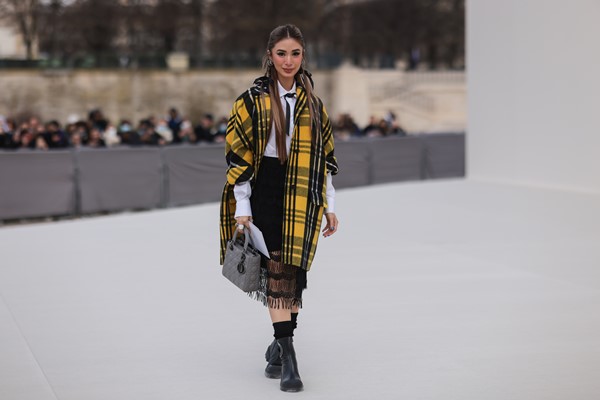 Mulher branca, com cabelo liso longo, posando para foto na porta do desfile da Dior, em Paris. Ela usa brincos de prata, camisa branca, gravata de fita preta, casaco longo com estampa xadrez preto/amarelo/branco da Dior, saia midi listrada preta com estampa de renda, anéis grandes de prata e strass.