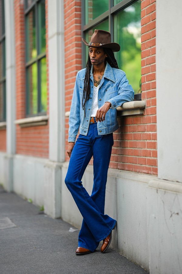 Homem negro, com tranças longas, posando para foto na Semana de Moda de Milão. Ele usa um chapéu de cowboy marrom com uma pena, uma jaqueta jeans azul, um pulôver branco de lã trançada com decote em V, um colar de pingente de corrente de ouro, um cinto de couro azul e camelo, calça jeans azul marinho e couro marrom brilhante botins de salto bloco.