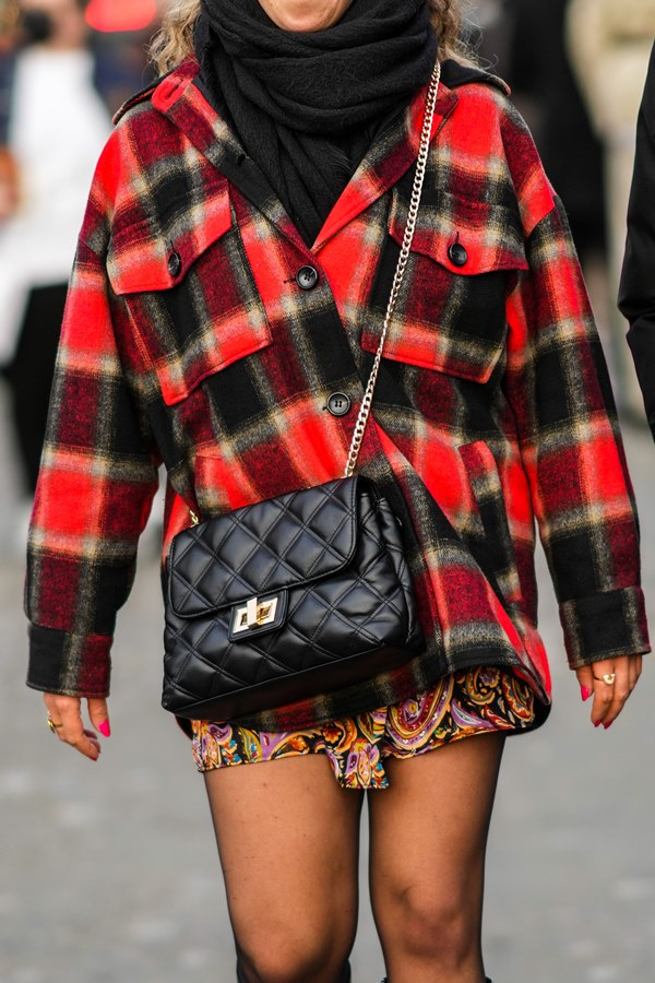 Mulher branca andando pela rua. Ela veste um lenço de lã preta, uma camisa xadrez vermelha e preta, uma bolsa crossbody acolchoada de couro preto brilhante, um vestido preto com estampa amarela / roxa / vermelha, meia-calça preta.