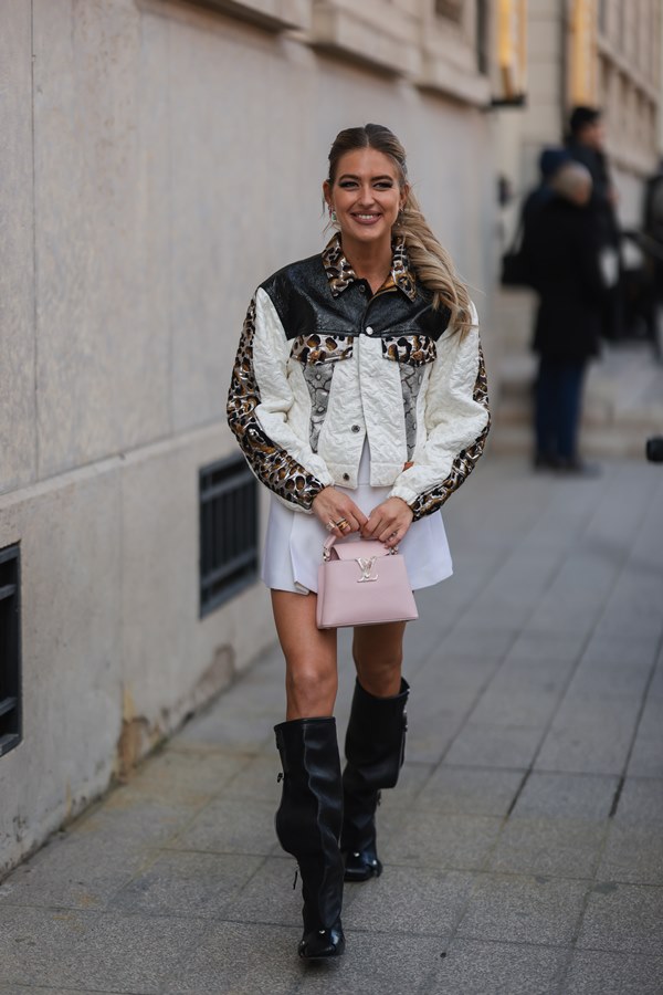 Mulher branca, com cabelo louros longos e ondulados, andando nas ruas de Paris. Ela veste uma camiseta branca, um padrão em relevo branco / jaqueta bomber de material misto com estampa de leopardo prata e marrom da Louis Vuitton, uma saia plissada branca, uma bolsa de couro rosa claro brilhante da Louis Vuitton,