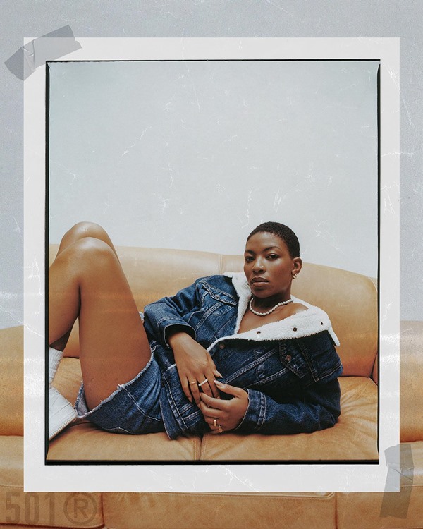 Modelo negra, com cabelo raspado, posando para campanha da marca Levi's. Ela usa uma jaqueta e um shorts jeans e está sentada em um sofá de couro marrom