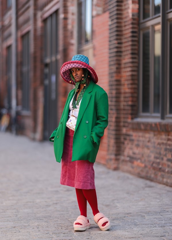 Mulher negra posando para foto nas ruas de Berlim, na Alemanha, e usando roupas estilosas e inusitadas. Ela veste um chapéu colorido da Gucci, jaqueta verde, saia rosa, bolsa transpassada branca da Louis Vuitton, meia calça vermelha com uma sandália branca de pelos.