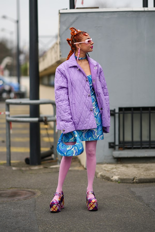 Mulher branca, com cabelo laranja em um penteado de trança, posando para foto nas ruas de Paris, durante a semana de moda na cidade. Ela usa brincos grandes e óculos de sol, tudo rosa; um vestido curto com estampa psicodélica nas cores brancas e azul; a bolsa na mesma estampa do vestido; meia calça rosa e uma sandália de salto xadrez.